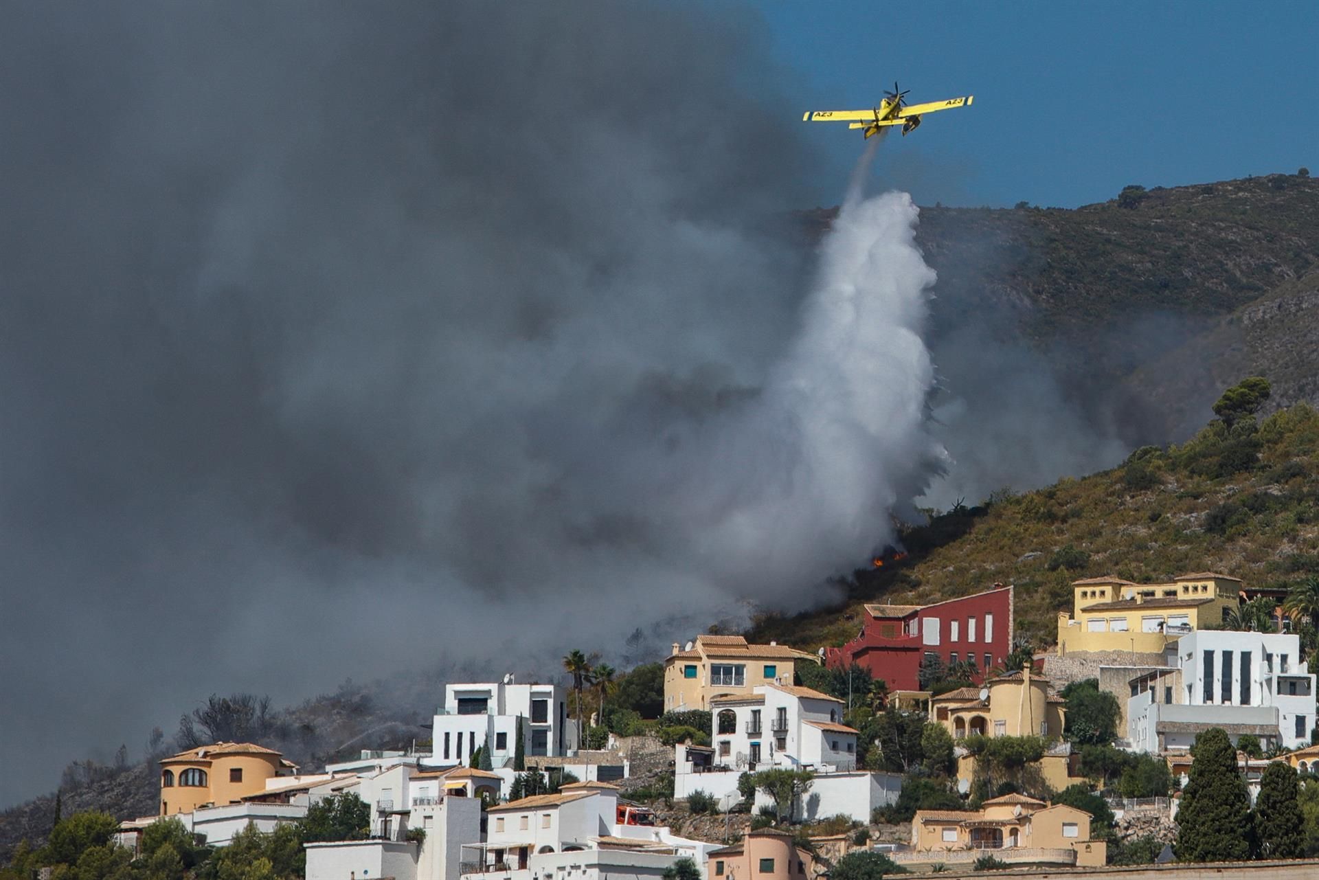En la imagen una avioneta realiza una descarga de agua sobre las llamas que avanzan hacia una zona de viviendas en la Atzubia